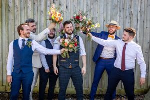 Groomsmen playfully hold up colorful bridal bouquets while posing with the groom against a wooden fence.