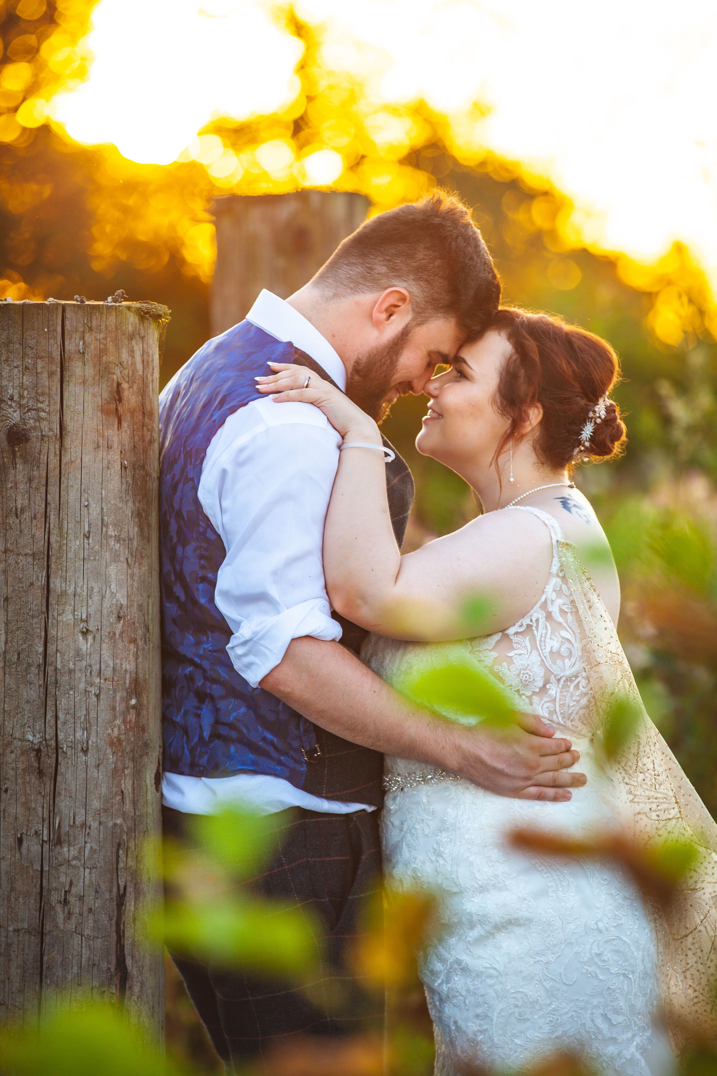 Bride and groom share a romantic golden hour moment embracing near rustic wooden posts in a vineyard.