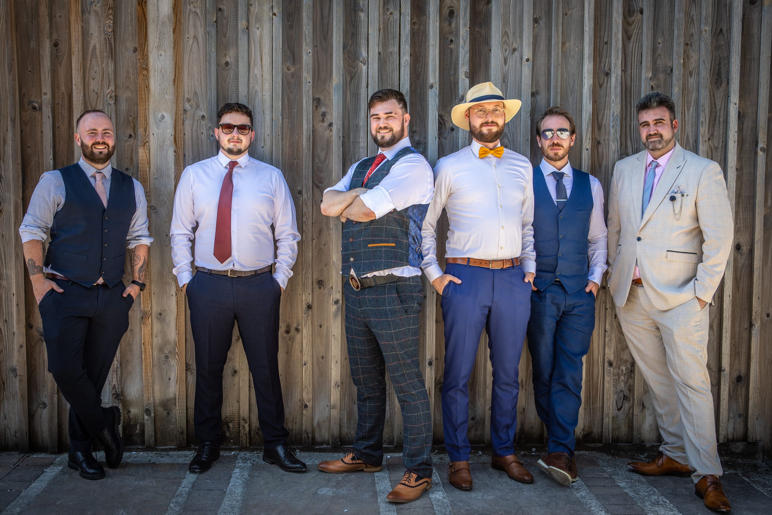 Six groomsmen in stylish suits and waistcoats pose confidently against a rustic wooden barn wall.