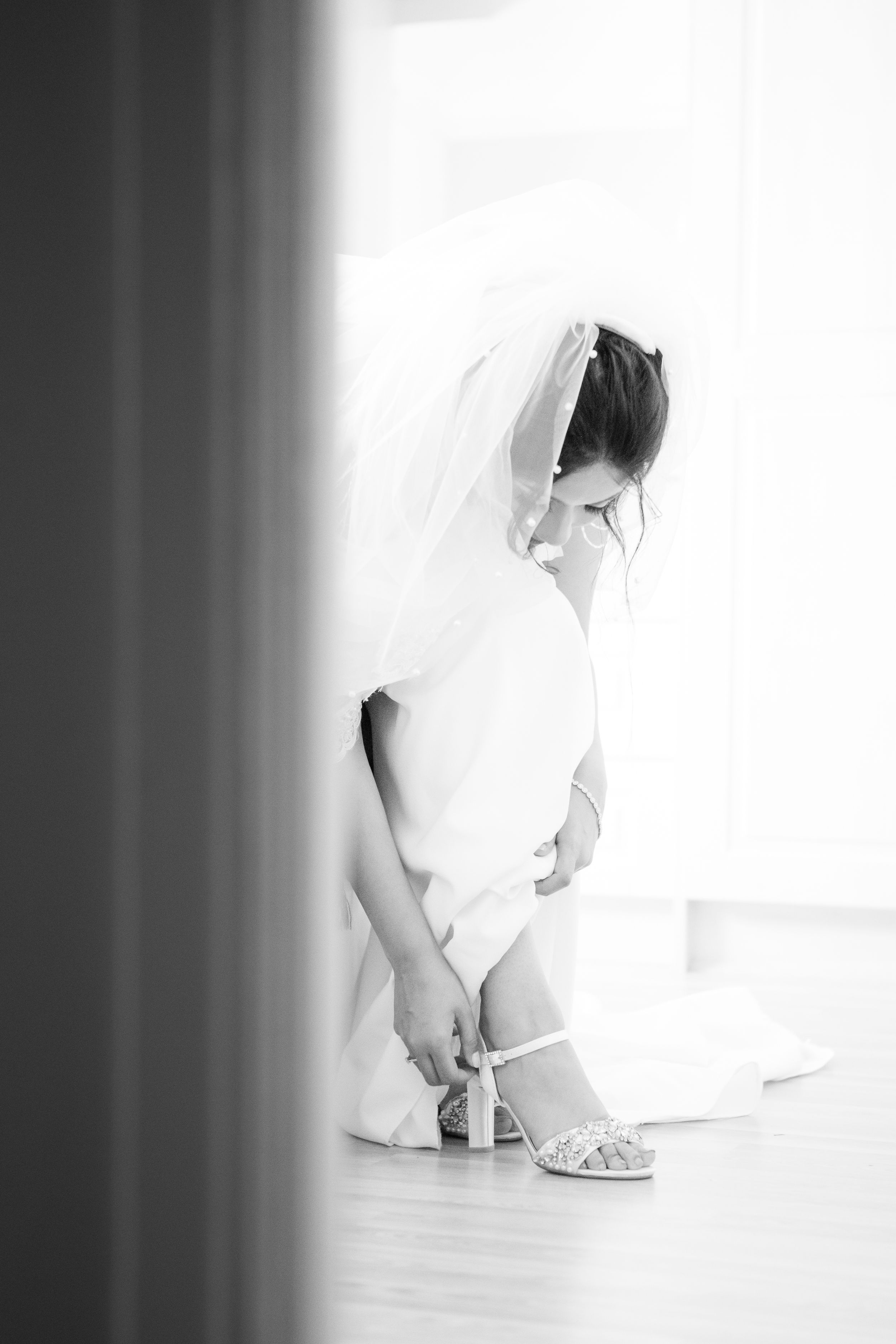 Black and white photo of a bride in a veil carefully fastening her strappy wedding heels.