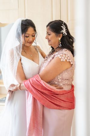 A mother tenderly adjusts her daughters wedding veil as the bride smiles softly in a bright room.