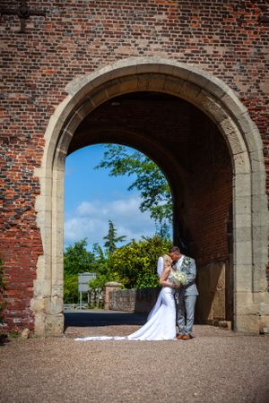 Bride and groom kissing under a historic brick archway with blue sky and green trees in the background.