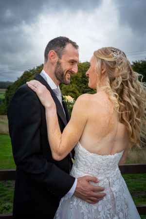 Bride and groom sharing a joyful glance outdoors with a cloudy sky and green countryside behind them.