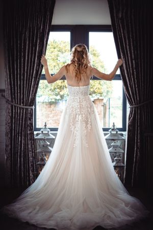 Bride in a stunning lace wedding gown standing at a large window holding open dark curtains.