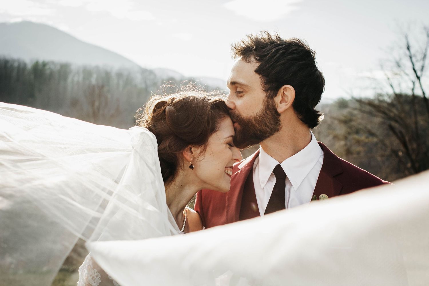 Newlywed couple embracing outdoors, groom in a red suit kissing the bride's forehead, her face partially obscured by a flowing veil.