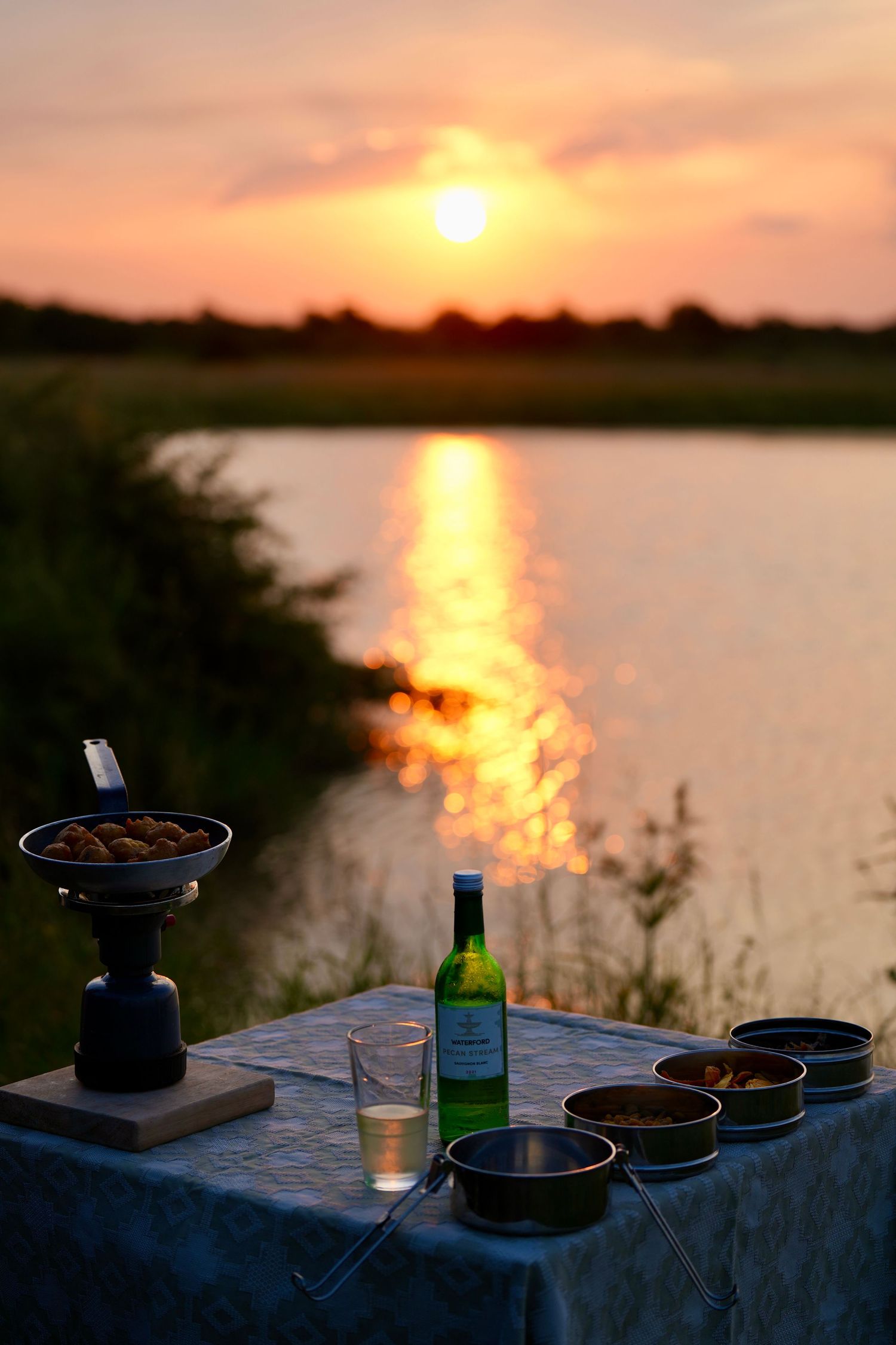 Sunset drinks and snacks arranged on a table by a waterfront with orange sky reflections.