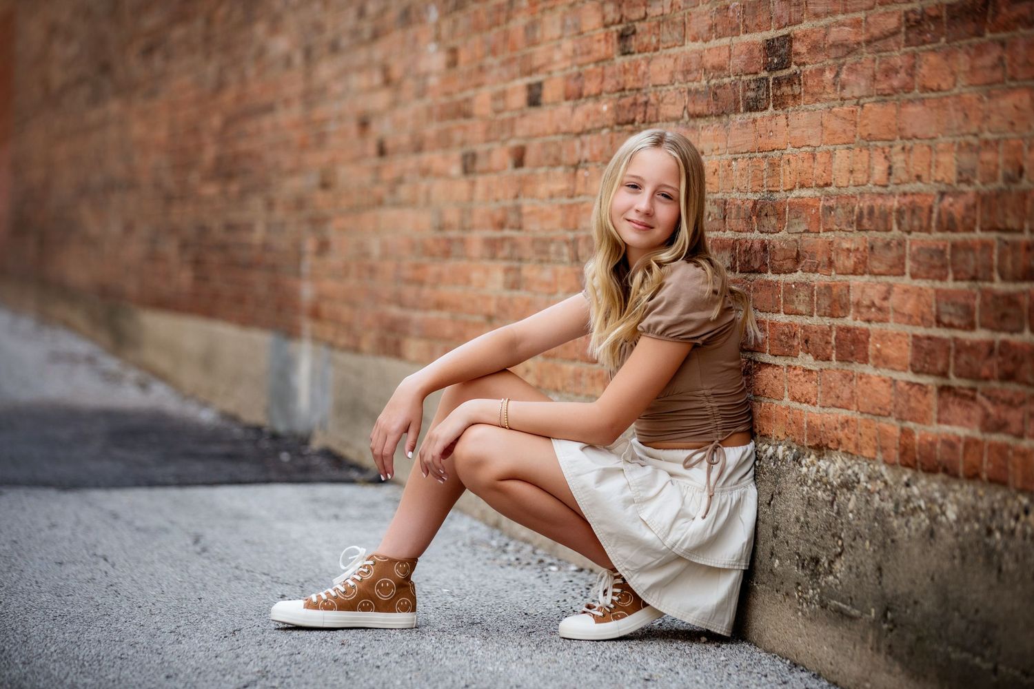 Young person in casual attire sitting against a brick wall outdoors.
