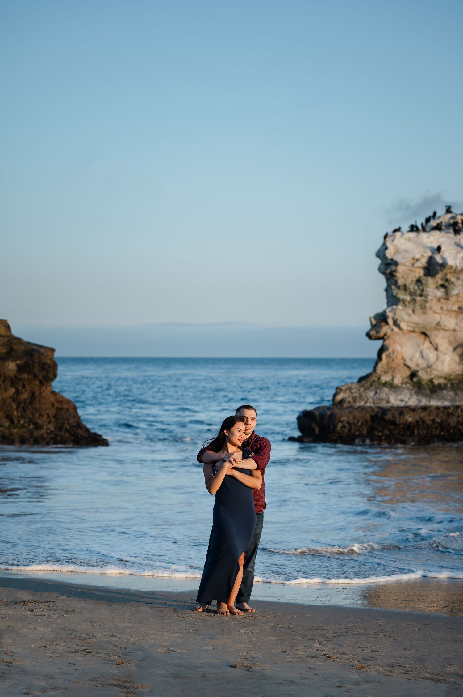A couple shares an intimate moment on a beach with dramatic rock formations in the background.