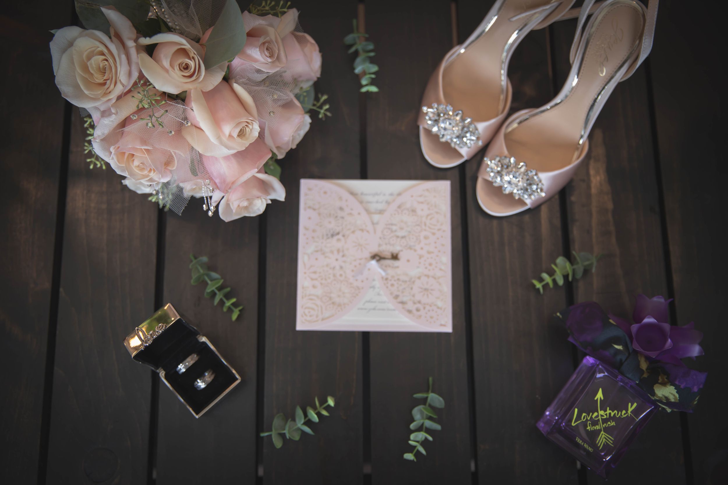 Wedding details with blush roses, elegant invitation, sparkly heels and wedding rings on dark wooden surface.
