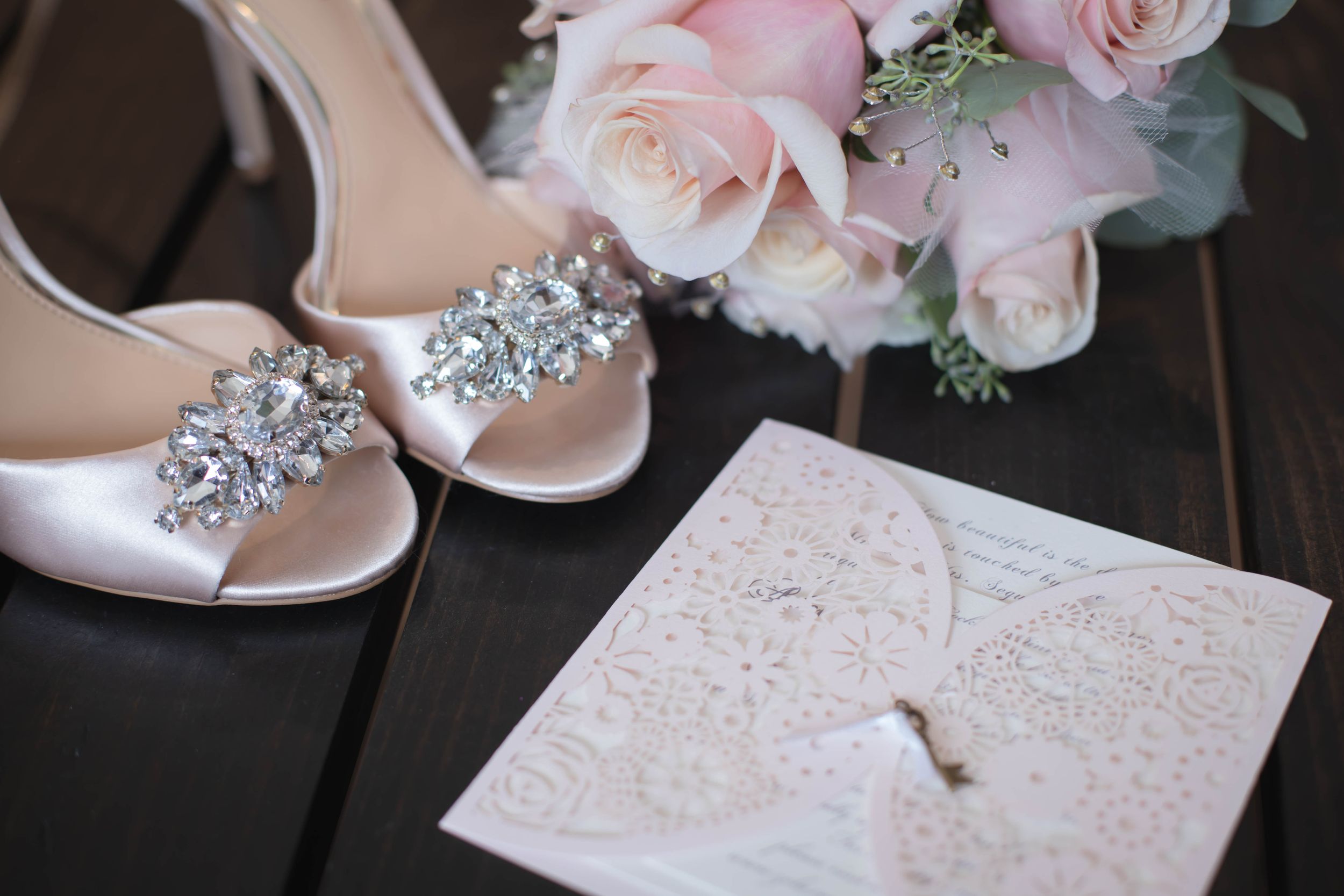 Close up of bridal shoes with crystal embellishments next to pink roses and white invitation.