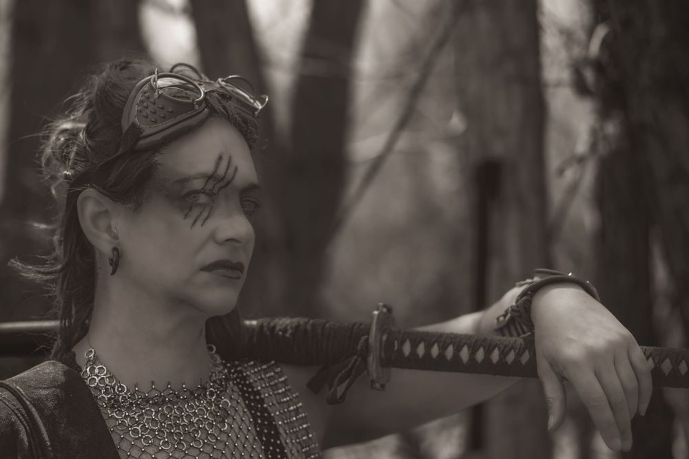 Dramatic black and white portrait with sword and ornate costume details in forest setting.