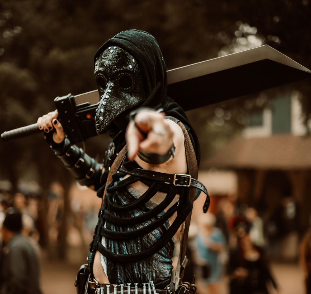 Cosplayer in dark post-apocalyptic costume with leather armor and face mask carrying a large prop weapon.