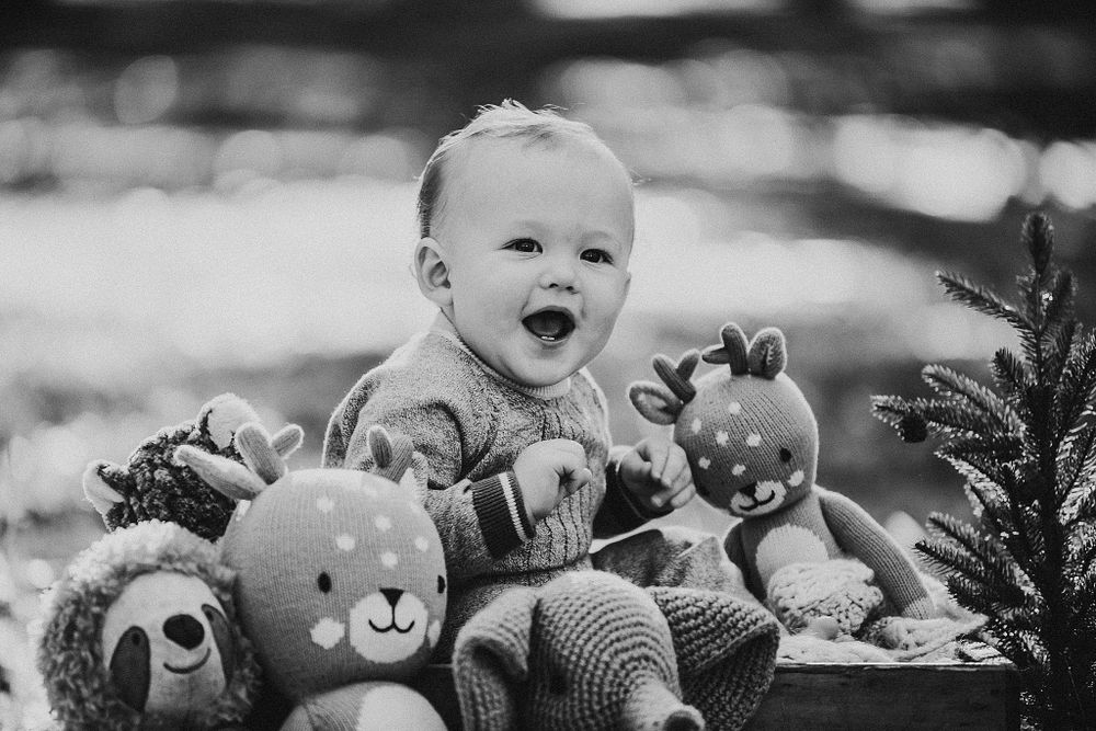 Happy baby sitting with collection of soft plush toys and stuffed animals in black and white portrait.