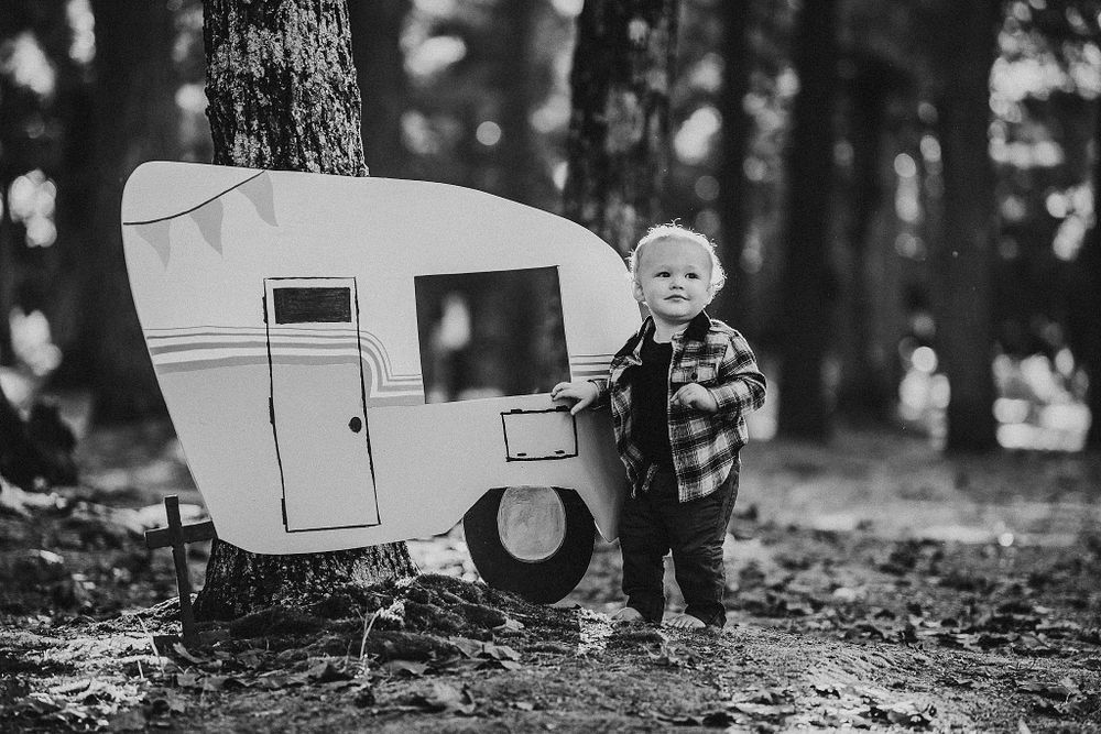 Black and white photo of small retro camper cutout display mounted on tree in woods.