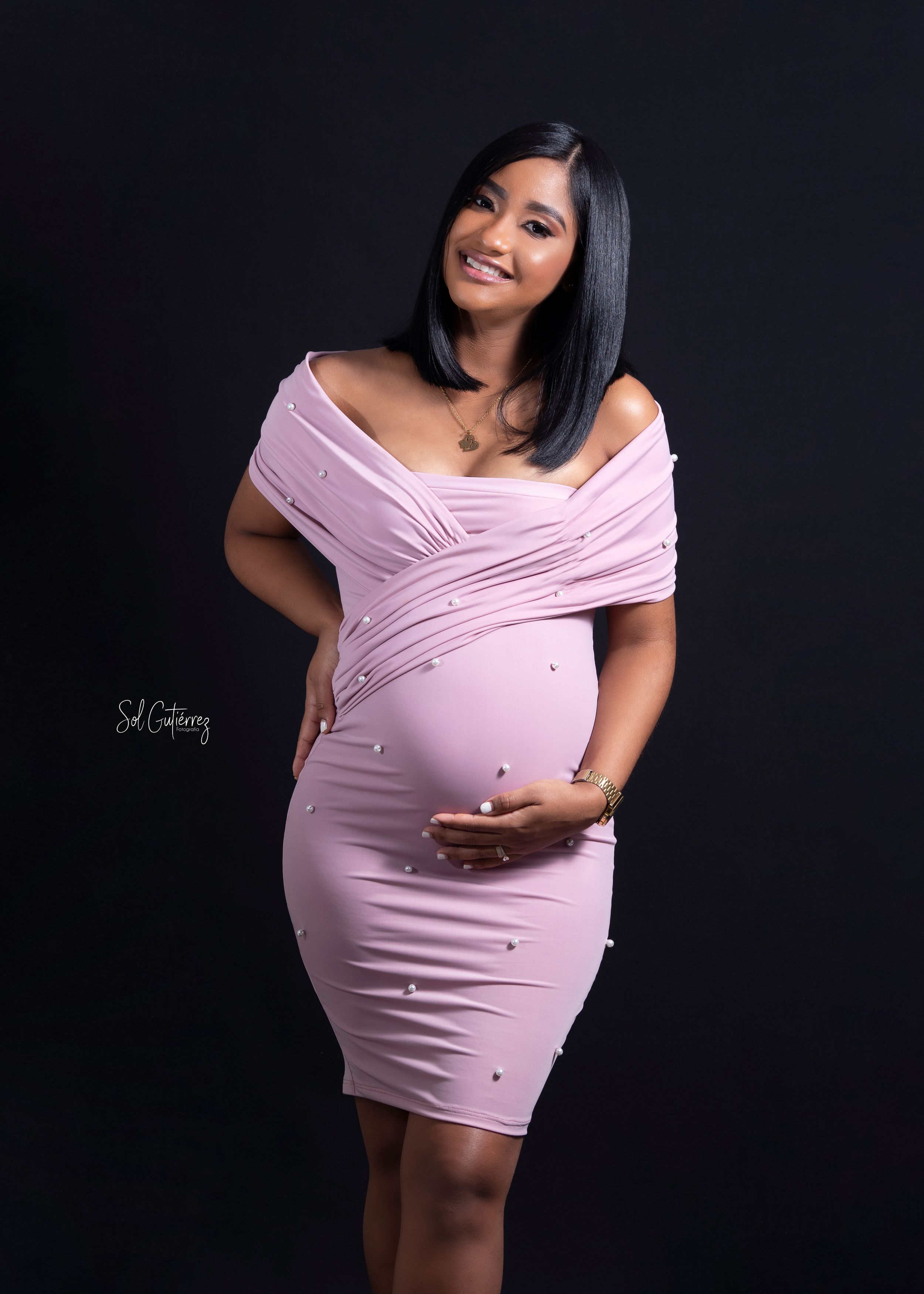 Studio maternity portrait in Lehigh Valley of smiling pregnant woman in elegant pink dress holding baby bump.