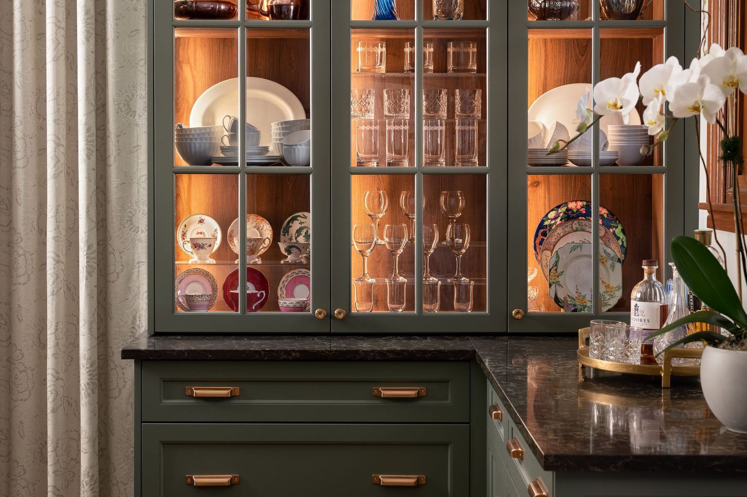 Illuminated glass cabinet displaying glassware collection with dark green cabinetry and marble countertop.