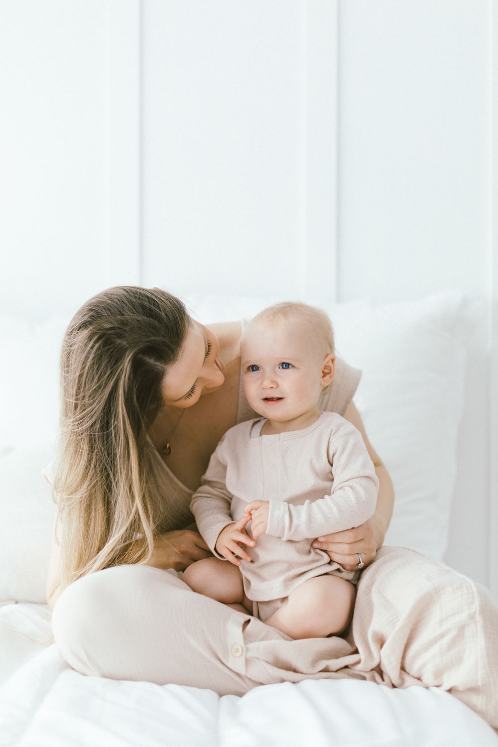 Sweet interaction between parent and baby in matching light colored outfits on white bedding.