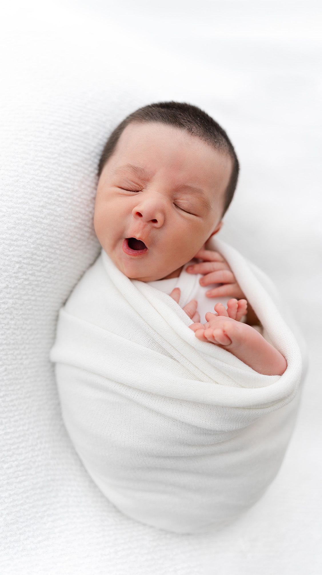 Sleeping newborn wrapped in soft white swaddle on textured background.