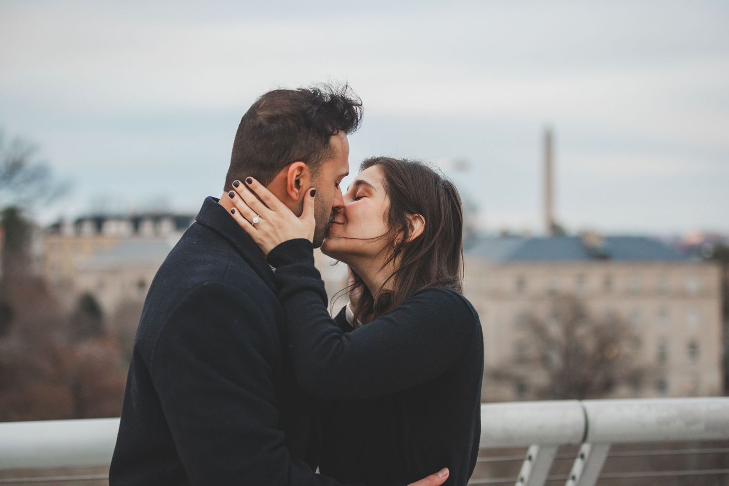 A romantic couple shares an intimate kiss on a balcony with a cityscape in the background.