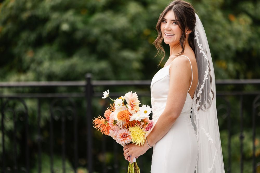 Brunette bride in sleeveless gown with lace veil holds a colorful dahlia bouquet outdoors.