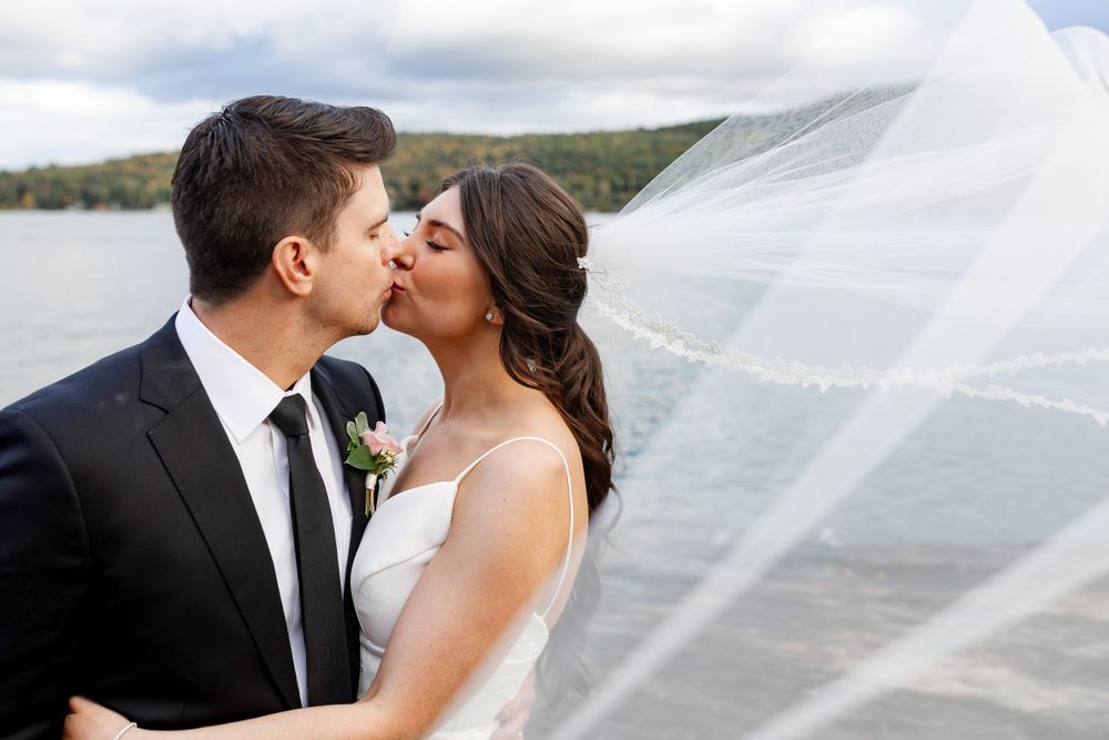 Bride and groom sharing romantic kiss under flowing veil with scenic lake and mountains in background.