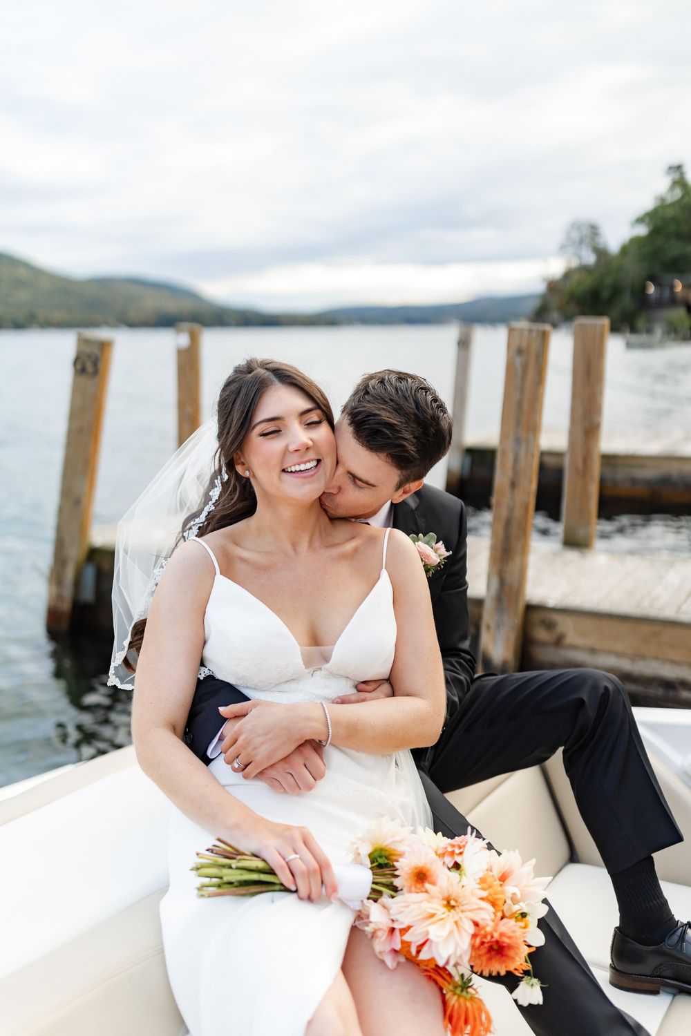 Groom kissing bride on neck while sitting on boat at lakeside dock, bride holding orange floral bouquet.