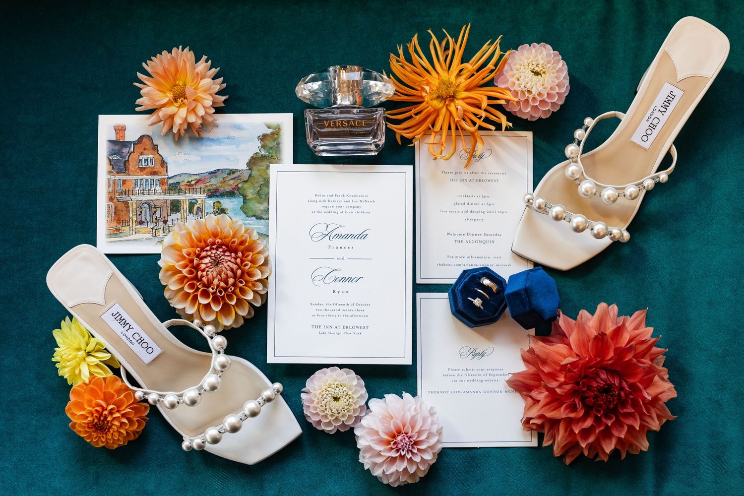 Wedding flat lay featuring pearl heels, dahlia flowers, invitations, and perfume on emerald velvet.