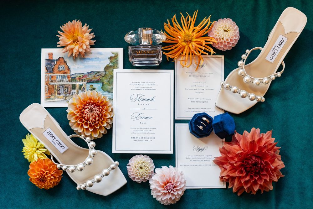 Wedding flat lay featuring pearl heels, dahlia flowers, invitations, and perfume on emerald velvet.