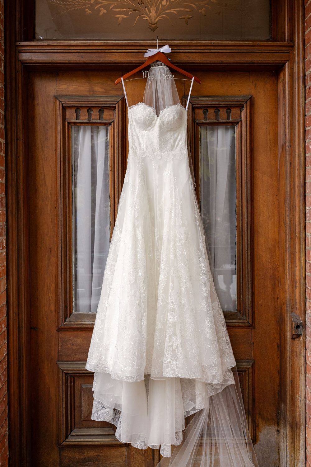 Elegant strapless lace wedding dress with veil hanging on ornate wooden door before the ceremony.