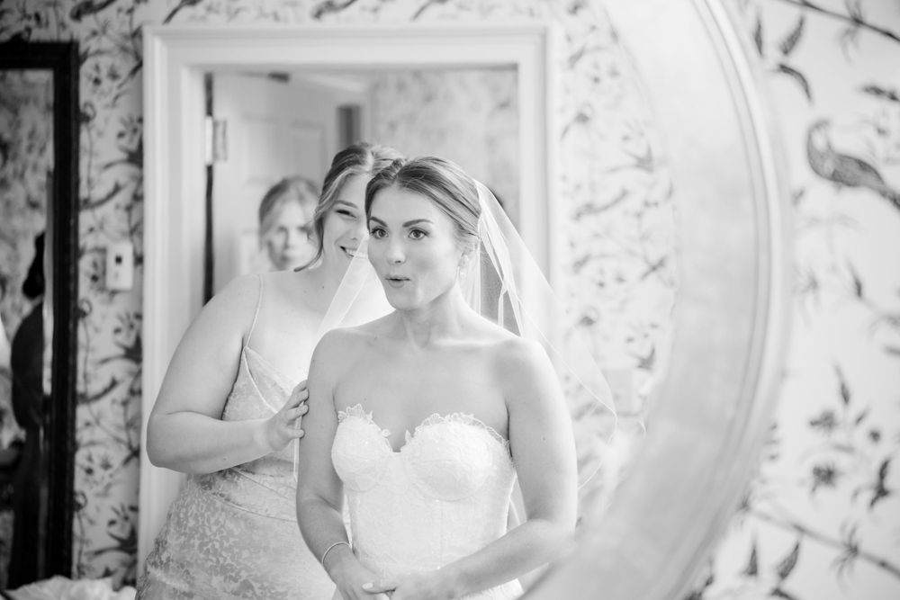 Black and white photo of bride in strapless lace gown with veil looking in mirror during preparation.