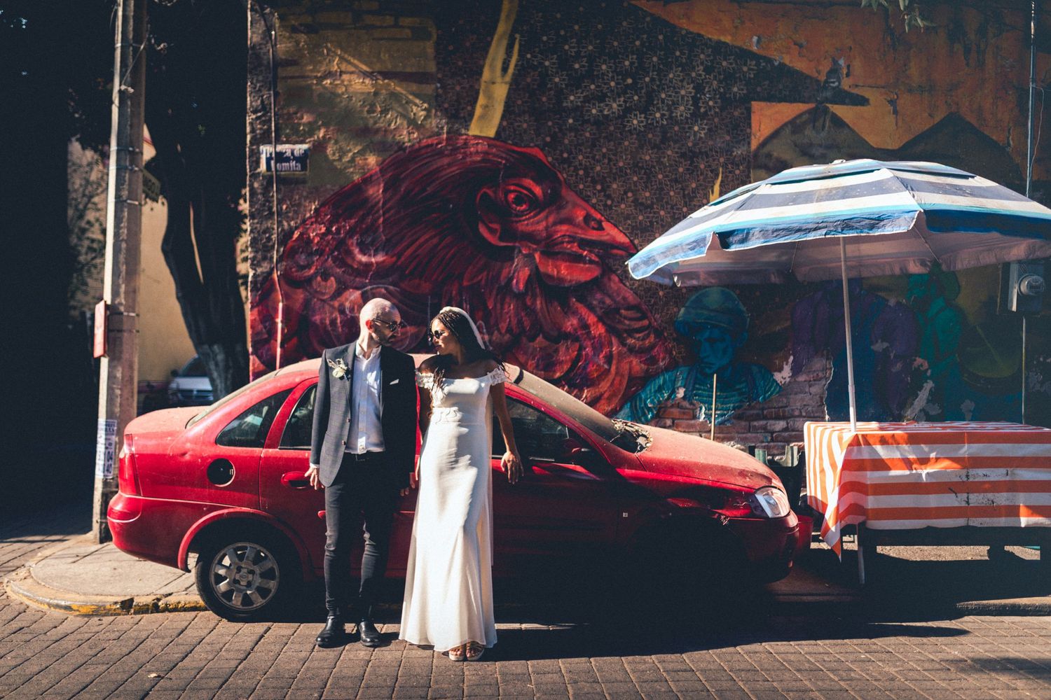 Newlyweds pose next to a red car with street art and umbrella in the background.