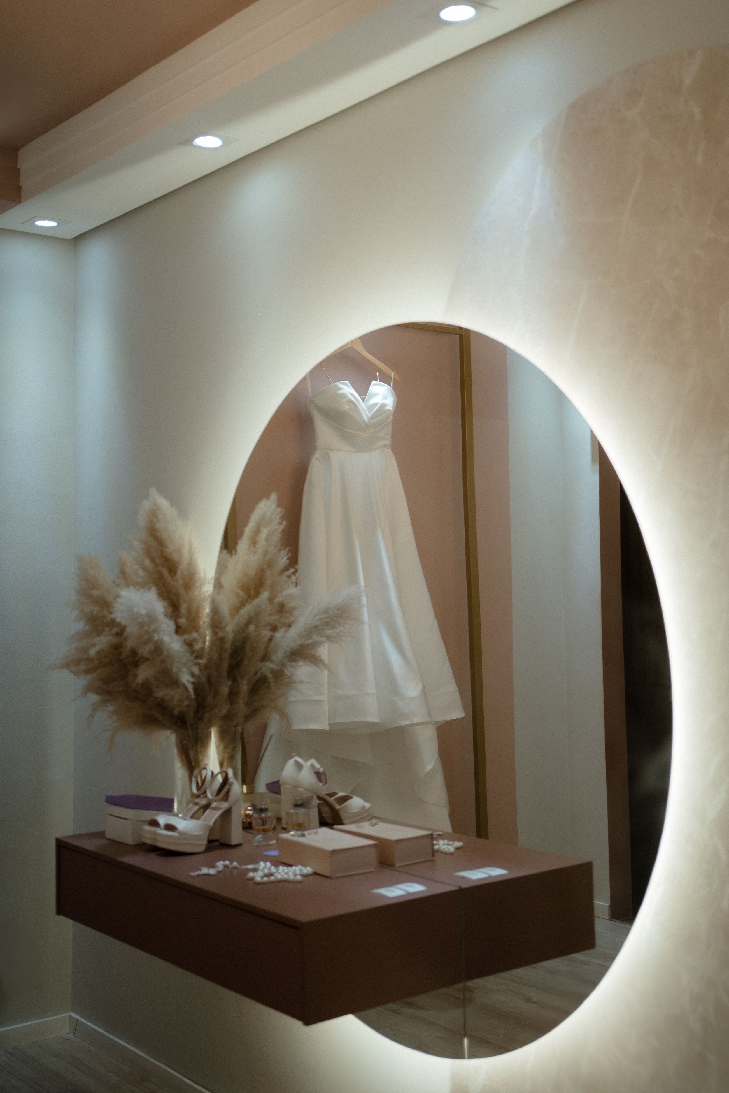 Bridal dress and accessories displayed on illuminated wooden shelf with pampas grass decor.