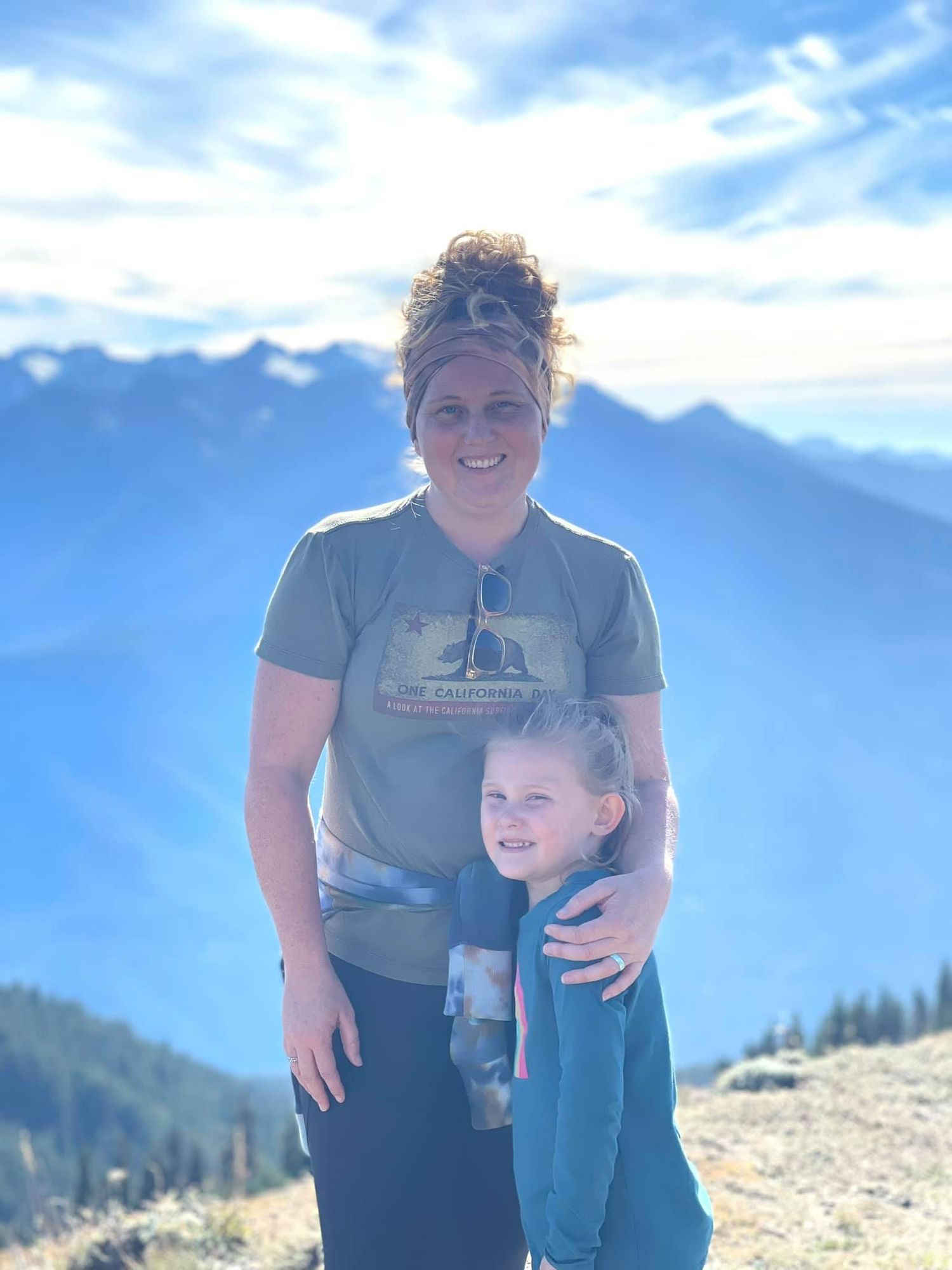 Two people stand together smiling on a mountain summit with blue skies and distant peaks in the background.