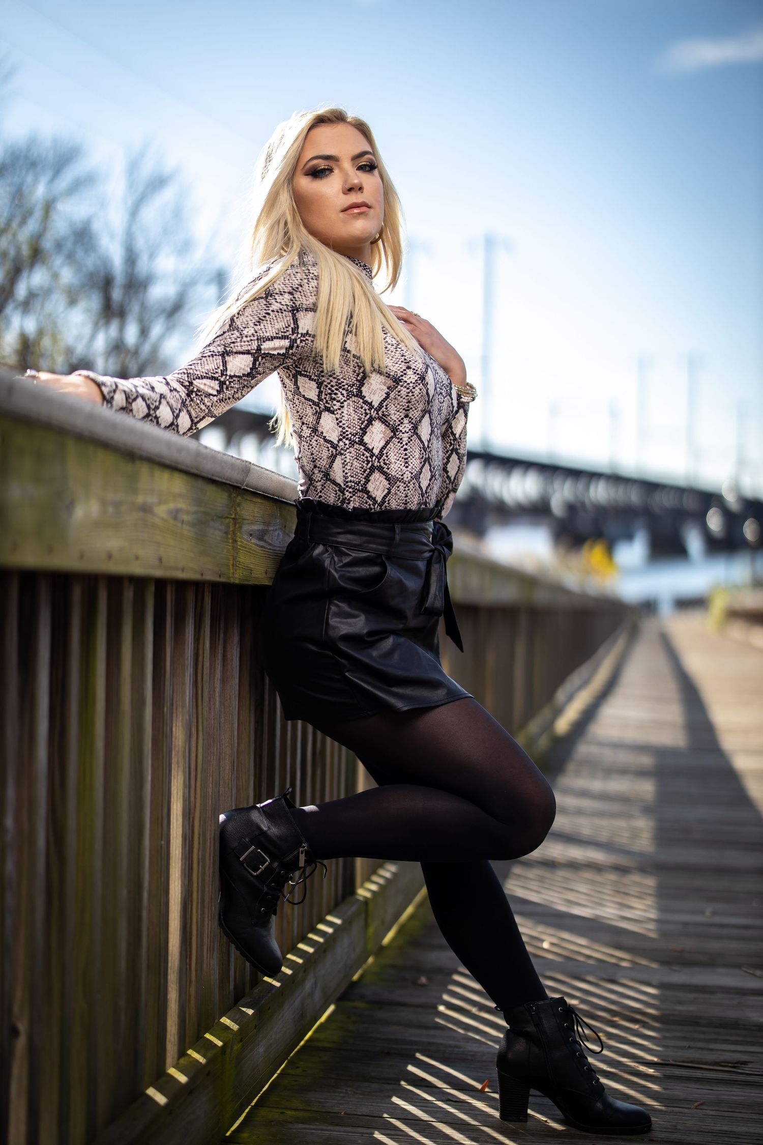 Fashion model poses in snakeskin print top and black skirt by bridge railing during outdoor photoshoot.