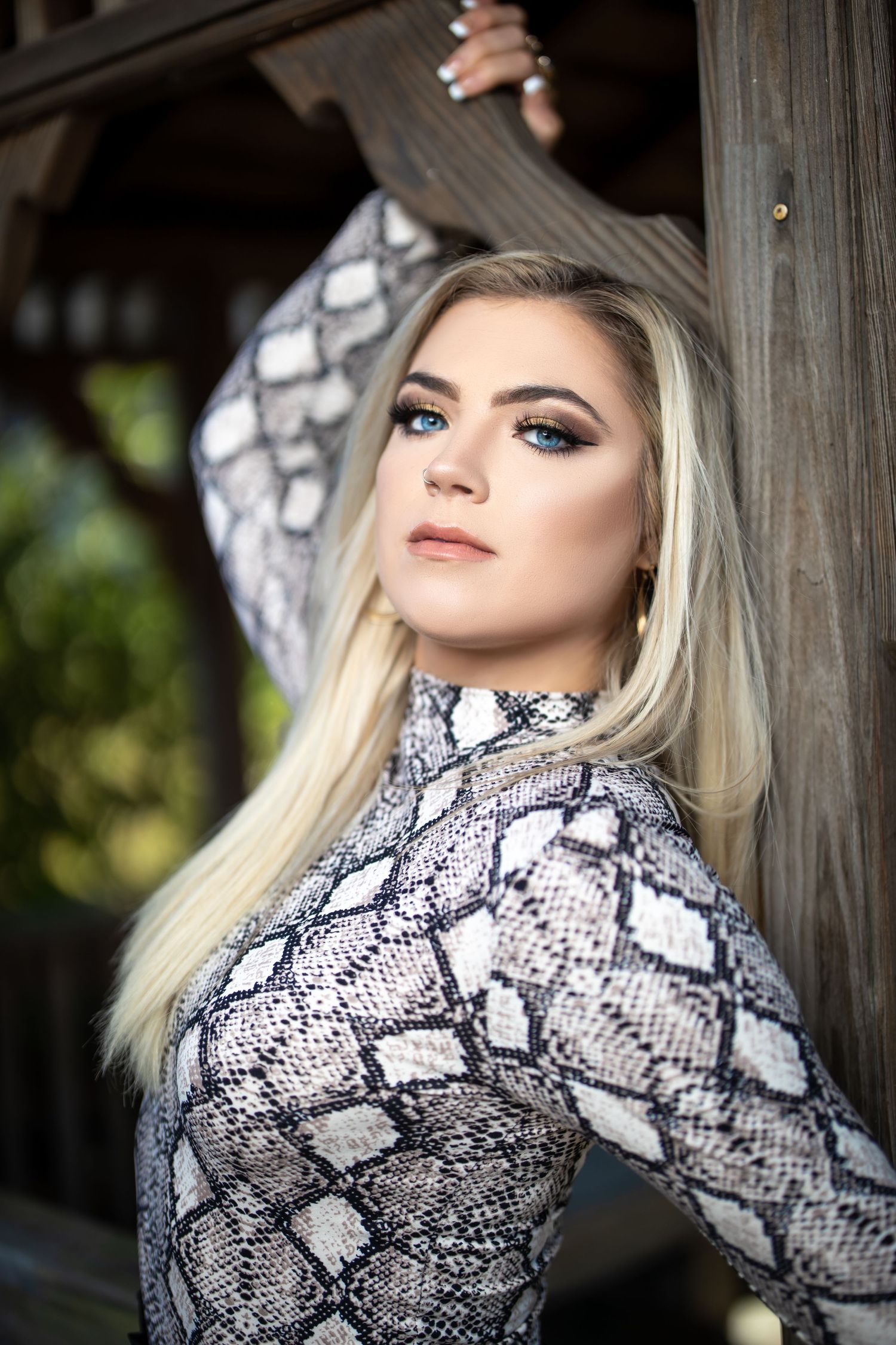 Close-up portrait in snakeskin print top with dramatic makeup against wooden gazebo.