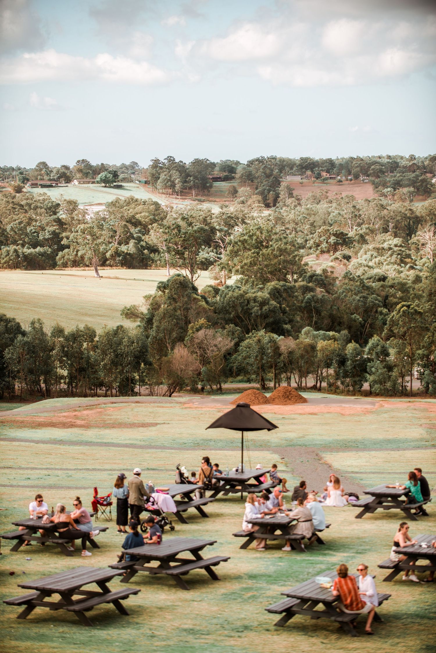 Outdoor picnic area with multiple wooden tables under an umbrella on a grassy hillside.