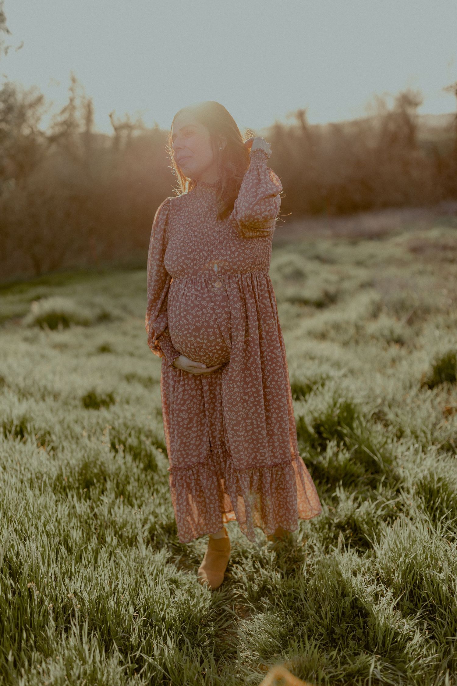 A pregnant woman in a floral dress stands in a sunlit field at golden hour.