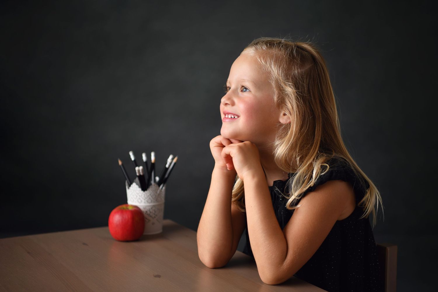 A student daydreams at a desk with an apple and pencil holder against a dark backdrop.
