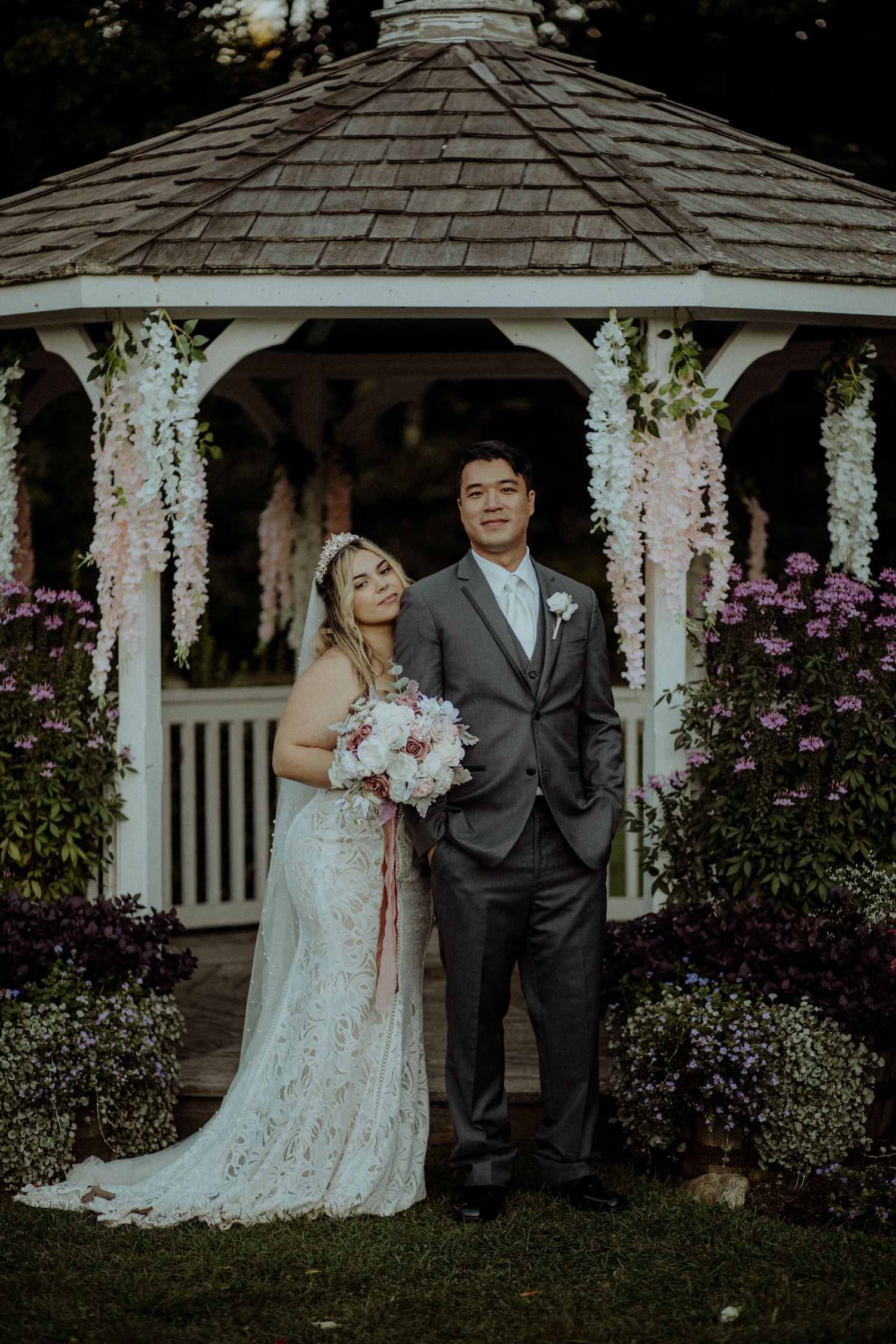 Bride and groom posing together under a floral-decorated white gazebo in a lush garden wedding setting.