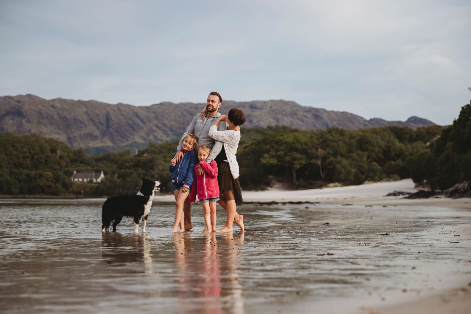 A family enjoys a playful moment on a beach with mountains in the background and their dog nearby.