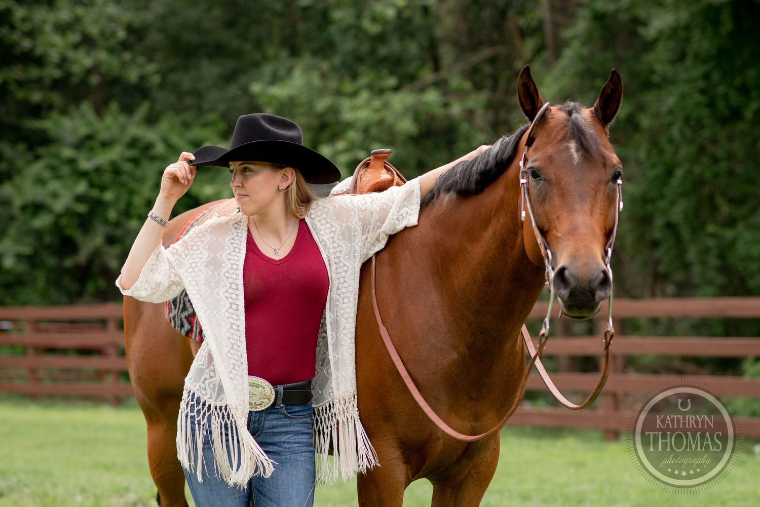 Rider in western wear and fringe jacket poses with chestnut horse against wooded background.