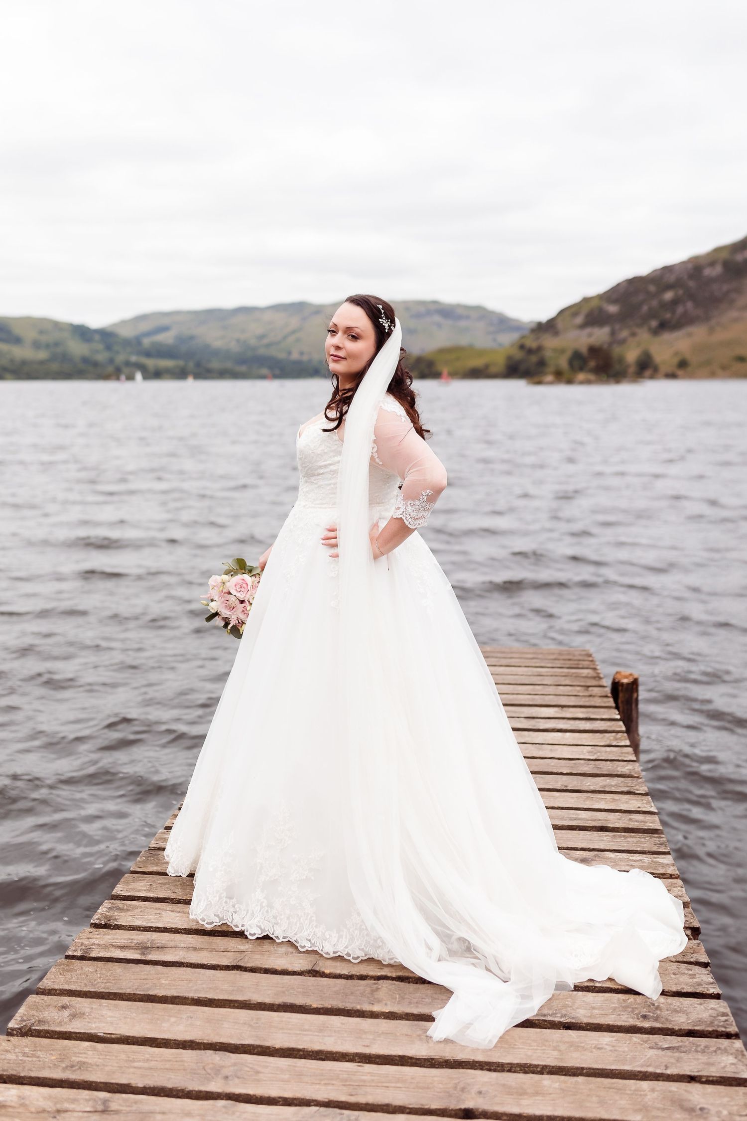 A radiant bride in a lace gown poses on a wooden pier overlooking a scenic lake.