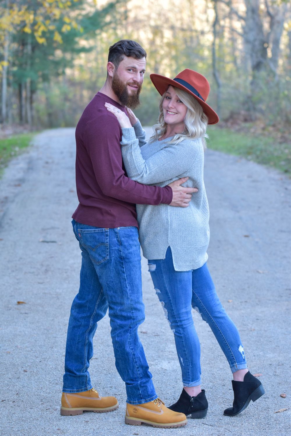 A couple embraces on a dirt path in autumn wearing casual sweaters and jeans.