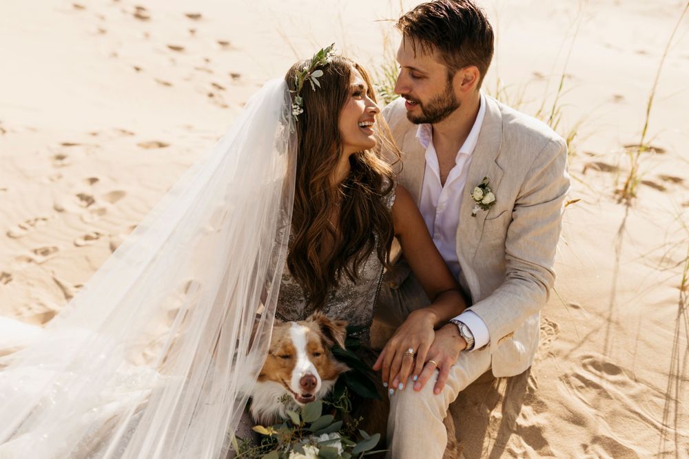 Bride in lace gown and floral crown gazes at groom while dog rests between them on sand dunes.