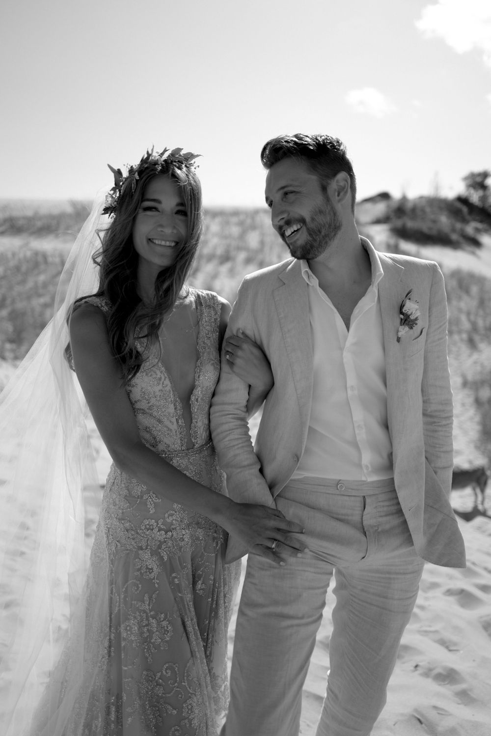 Black and white photo of smiling bride and groom standing together on sandy beach dunes.