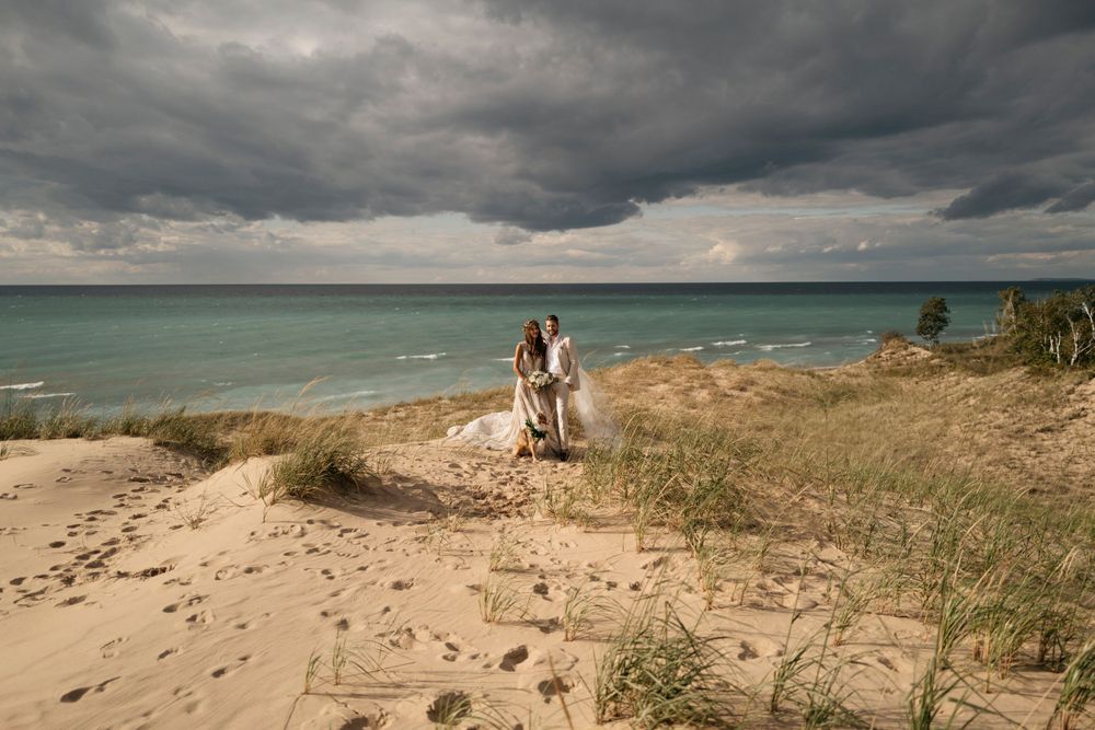 Bride and groom standing on sand dunes with dramatic stormy sky and lake in the background.