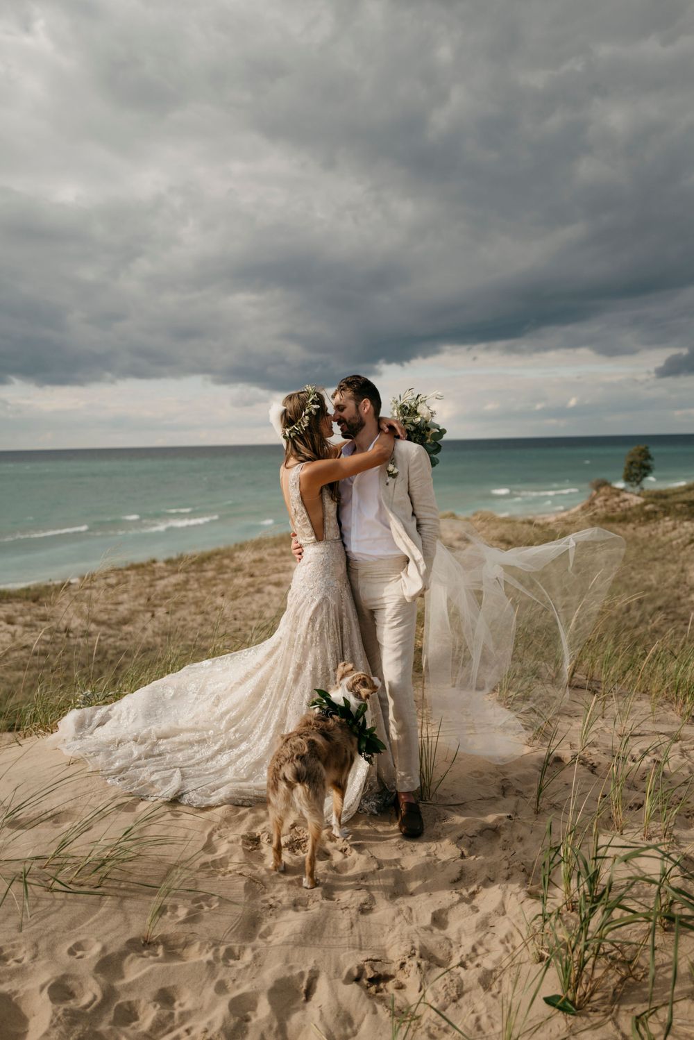Wedding couple with dog on sand dunes, bride in lace gown with flowing veil oceanside.