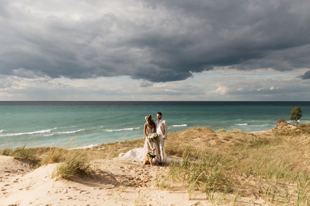 Bride and groom stand on sandy dunes overlooking turquoise lake under dramatic stormy sky.