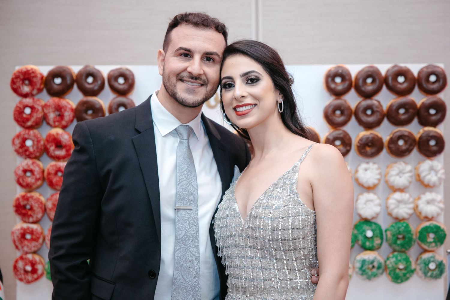 A couple poses in formal attire in front of a decorative donut wall display.