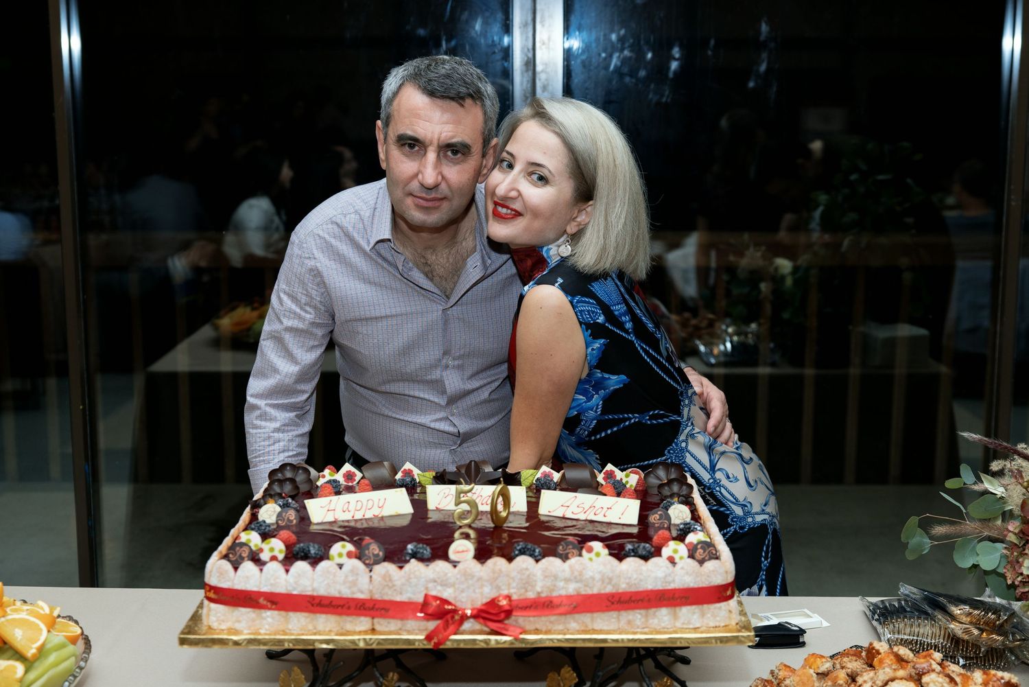 A couple poses behind a decorated birthday cake at an evening celebration.